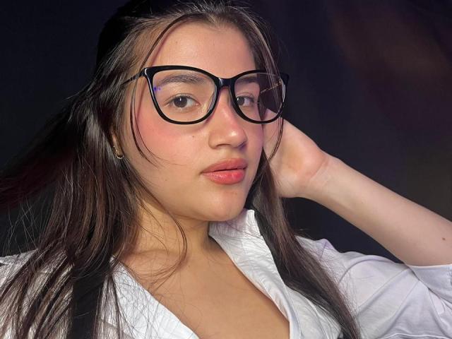 IrisBlair with black glasses, pinkish cheeks, and pink lips, wearing a white shirt, hand in hair, dark straight hair, soft skin, and a slightly serious expression.
