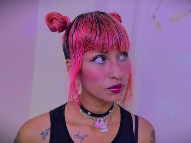 MilaRouge has pink and black double buns, bright pink lipstick, a black choker with a skull pendant, and tattoos on her chest and arms. Her pink hair is vibrant.