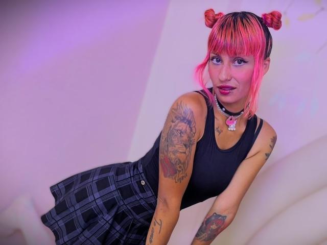 MilaRouge with pink hair in double buns, black tank top, and plaid skirt, showing off tattoos on arms and chest, wearing a choker with a pink heart.