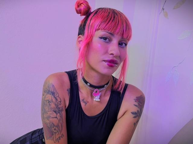 MilaRouge with pink hair in buns, black tank top, choker with pink gem, tattoos on arms, purple eyeshadow, pink lipstick, and a cute face, sitting.