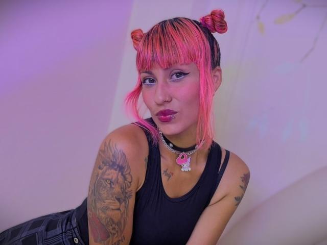 MilaRouge with pink hair in buns, black tank top, choker, tattooed arm, pink lipstick, and plump lips, leaning forward, showing off her tits, looking sexy and cute.