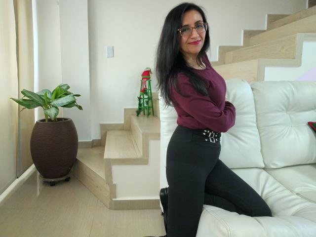 Cyntiia with long black hair, glasses, red lipstick, maroon sweater, black high-waisted pants, sitting on white leather couch, wooden stairs, green plant, brown pot, red ornament.