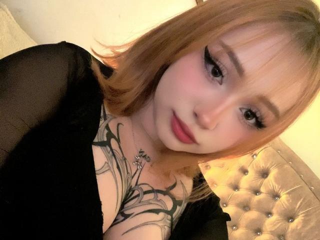 DarkCourtney with short blonde hair, black eyeliner, pink lipstick, and black top showing her tits and tattoo on her chest. She's looking at the camera with a slight smile.