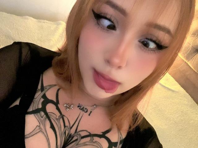 DarkCourtney with light skin, red hair, and black eyeliner, looking up with pink lips, black top, and large tattoos covering her tits. Her nose is pierced.
