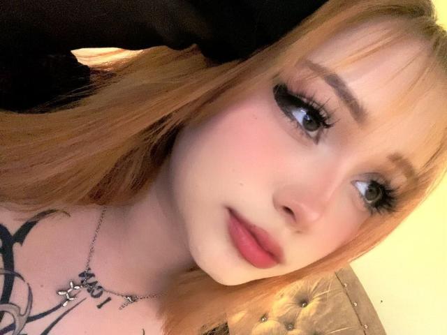 DarkCourtney has red hair, fair skin, and long eyelashes. She's looking at the camera with pink lips and a small tattoo on her chest. She's wearing a black top.