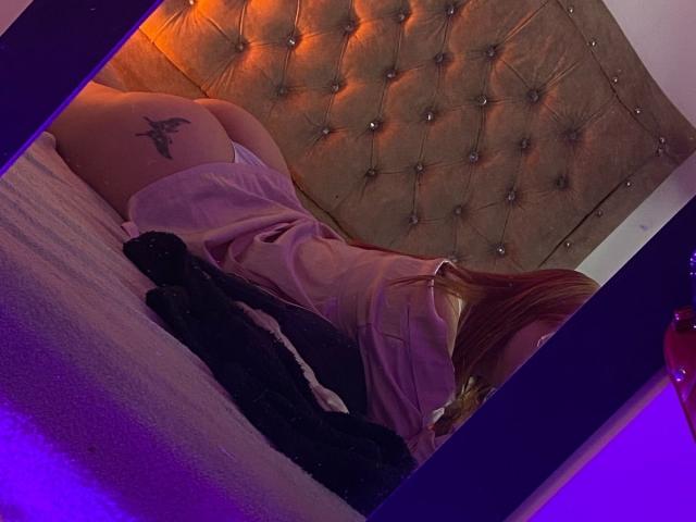 DarkCourtney, naked from the waist down, on a bed with a tufted headboard, showing a black tattoo on her ass, wearing a pink shirt, in a mirror selfie.
