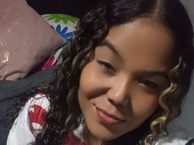 Estrellya has curly black and blonde hair, a warm brown skin tone, and a slight smile, with subtle makeup, lying down with a pink pillow and floral blanket.