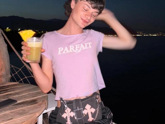 OnlyMollyG with short brown hair, pink crop top, and black belt with pink crosses, holding a yellow drink with a lemon slice, standing on a wooden dock at night.