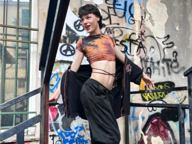 OnlyMollyG with short dark hair, wearing an orange tie-dye crop top and black pants, standing on a graffiti-covered stairway, jacket open, showing a slim, flat-assed, small-titted, midriff.