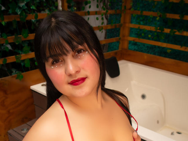 CataleyaHankl with dark hair, red lipstick, and freckles, wearing a red strap, sitting in a hot tub, smiling at the camera. Her tits are partially visible.
