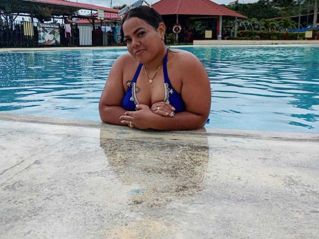 Rizantella, a chubby woman with brown skin and black hair, in a blue bikini, showing her big boobs, leans on a pool edge, water up to her chest.