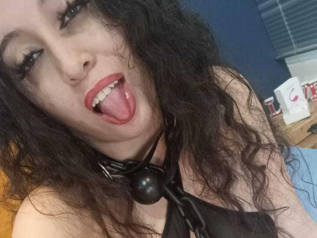 KendraSimons with long, curly black hair, black leather choker with ball, and open mouth showing tongue, has pink lipstick and black eyeliner, looking directly at camera.