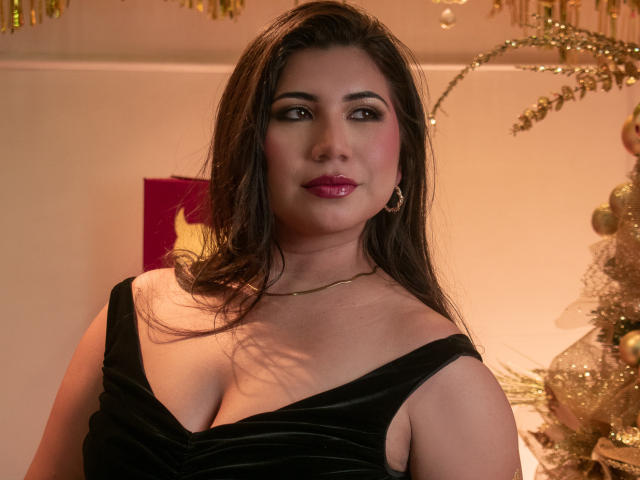BabeCloe has a medium build with a slightly curvy figure, wearing a black, off-shoulder velvet dress. She has long, wavy brown hair and is looking directly at the camera with a subtle smile. Her makeup is bold, with dark eyeliner and bright red lipstick. She's wearing small, gold hoop earrings. The background is softly lit with gold and pink decorations, including a glittery tree. The image has a warm, inviting tone.