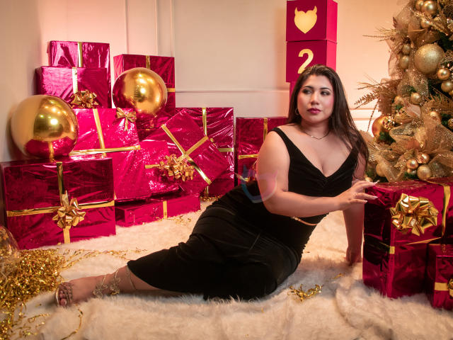 BabeCloe sits on a fluffy white rug, wearing a black, sleeveless, low-cut dress. She has long, dark hair and red lipstick. She's surrounded by shiny red and gold-wrapped gifts, gold balloons, and a glittery Christmas tree. Her right hand touches a gift, while her left arm rests on the rug. The room is warmly lit with a heart-shaped decoration on a wall in the background. The image has a festive, warm, and luxurious feel.