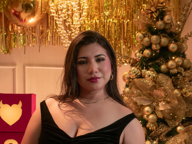 BabeCloe stands in a festive, gold-decorated setting. She has long, wet brown hair, wearing a black velvet dress with a deep neckline, showing cleavage. Her makeup is bold with pink lipstick. A gold Christmas tree adorned with glittery ornaments and gold ribbons is on her right. A pink gift box with a heart cutout is on her left. The lighting is warm, highlighting her fair skin and subtle blush. The overall composition is rich and celebratory.