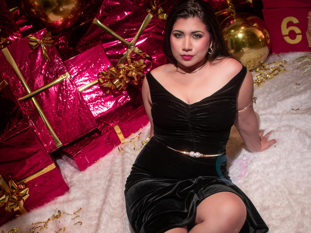 BabeCloe sits on a white, fluffy rug. She wears a black, off-shoulder velvet dress with a gold belt. Her dark hair is loose, and she has bold red lipstick. She's surrounded by pink and gold presents with shiny ribbons. Gold ornaments are scattered around. Her left arm rests on the rug, and her right hand is on her knee. The image is well-lit with a warm, festive glow. She has a confident, slightly serious expression. The composition is centered, emphasizing her and the vibrant, festive background.