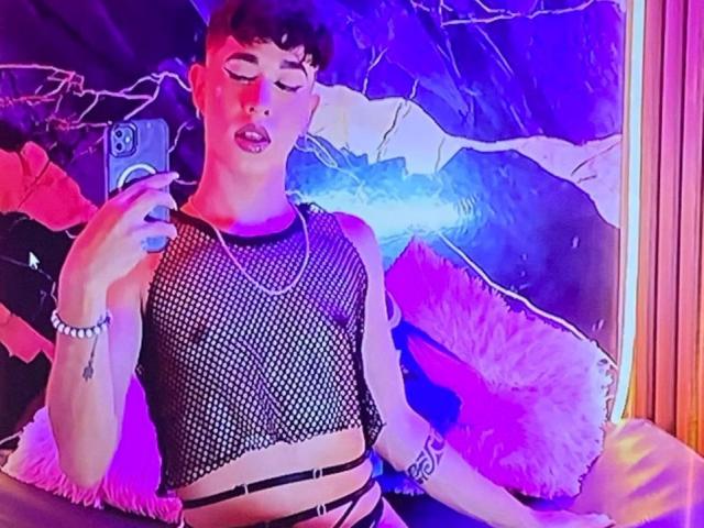 BrigitteLove with short dark hair, wearing a black fishnet top, holding a phone, purple and blue lights, white fluffy pillow, black strappy bottom, and a silver bracelet.