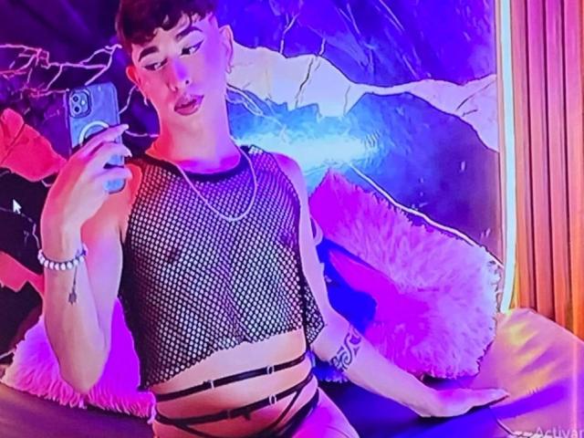 BriggitteLove with short curly hair, wearing a black fishnet top, black strappy underwear, and a silver chain, taking a selfie in a pink and purple lit room.