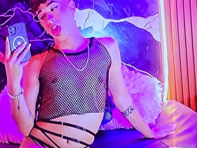 BriggitteLove wearing a black fishnet top, exposing her tits, with strappy black bottoms, taking a selfie with pink and blue lights, tongue out, and a tattoo on her arm.