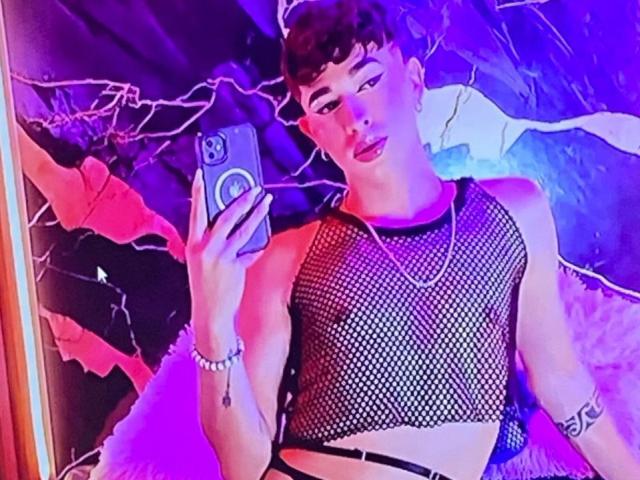 BriggitteLove takes a selfie in a mirror. They have a slim build, dark curly hair, and wear a black fishnet top revealing a toned torso. They hold a smartphone with a circular button in the center. BriggitteLove has pink lipstick, a silver bracelet, and a tattoo on their right forearm. The background is a vivid, purple-blue pattern. The photo has a bright, neon glow. BriggitteLove looks at the mirror with a slightly pouty expression.