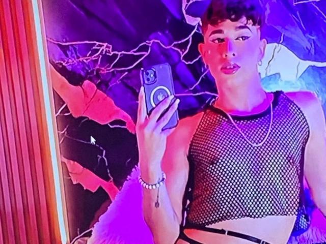 BriggitteLove with short curly brown hair, wearing a black fishnet top, holding a phone, looking in a mirror, purple and pink neon lighting, bracelet, and necklace.