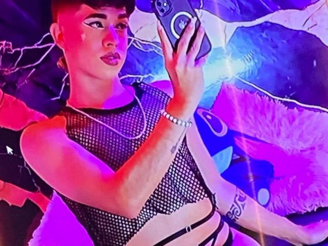 BriggiteLove with short dark hair, wearing a black mesh top, taking a selfie with a phone, has a tattoo on her right arm, and is wearing a silver bracelet.