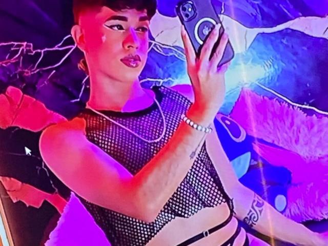 BriggitteLove with short brown hair, wearing a black mesh top, taking a selfie. She's got a silver bracelet, a necklace, and a tattoo on her right arm.