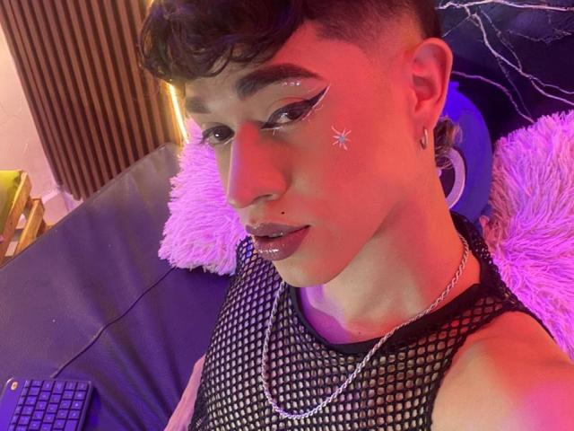 BriggiteLove with short curly brown hair, winged eyeliner, glitter star on cheek, shiny lips, fishnet top, necklace, purple and pink lighting, lying on blue chair with pink pillow.