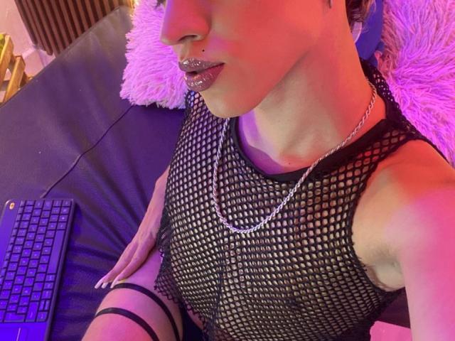 BriggitteLove with glittery lips, black fishnet top, silver chain, and strappy thigh, sitting on a bed with a laptop, pink and purple lighting.