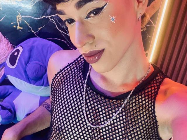 BrigitteLove has a pale skin tone, dark curly hair, and is wearing a black mesh sleeveless top. They have winged black eyeliner, a small silver star on their right cheek, and dark purple lipstick. A silver nose ring is visible on their right nostril. A silver chain necklace hangs around their neck. They are looking to the right with a slight smile. A purple plush toy with a large white eye is in the background. Bright, warm, and purple lighting creates contrasting highlights.