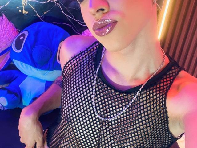 BriggitteLove with shiny lips, wearing a black fishnet top, silver chain, and no underwear, showing a slim, tan body with a cute, blue, stuffed animal to the left.
