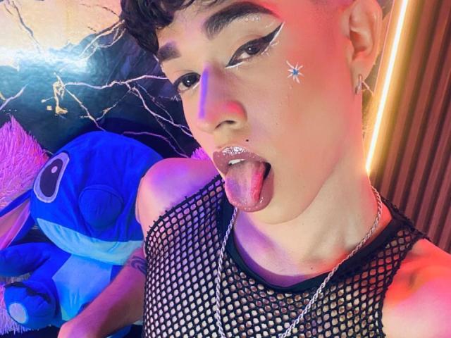 BriggitteLove has shiny makeup, star on cheek, black fishnet top, tongue out, piercing, and short curly hair, with neon pink and blue lighting. Visible ass tattoo on left.