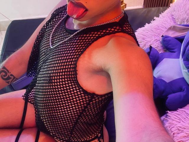 BriggitteLove is seated on a dark sofa, wearing a black fishnet bodysuit that reveals their pale skin. Their tongue is extended out, touching the side of their mouth. They have a tattoo on their left forearm and are wearing a silver chain necklace. A purple and blue plush toy is on the right. The lighting is pink and orange, creating a vivid, almost neon effect. The fishnet bodysuit is tight, highlighting their slender build. The image is a close-up, focusing on BriggitteLove's upper body and face.