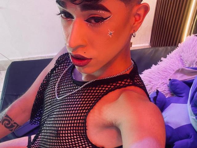 BriggitteLove with short brown hair, makeup, star tattoo, black fishnet top, earrings, red lipstick, arm tattoo, and chain necklace, looking at camera, sitting on dark couch.