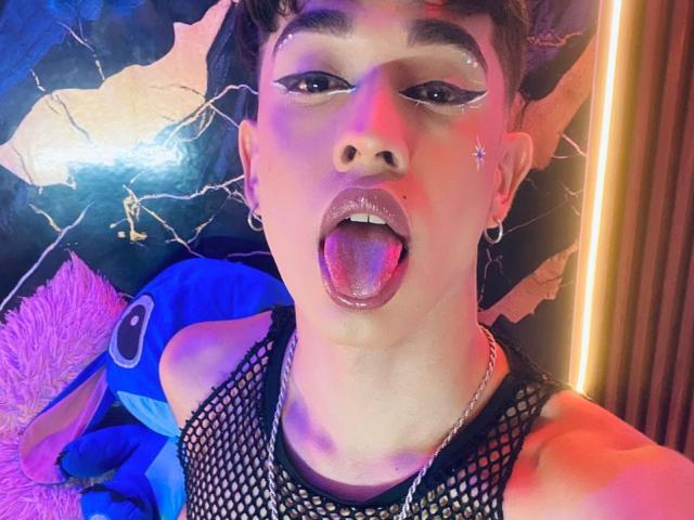 BriggitteLove has short, curly brown hair and is wearing dramatic black eyeliner with winged tips. Their tongue is out, coated in saliva. They have small star-shaped glitter on each cheek. BriggitteLove is wearing a black fishnet top and silver earrings. The photo has a mix of purple, pink, and blue lighting with a textured dark background. The overall style is bold and edgy.