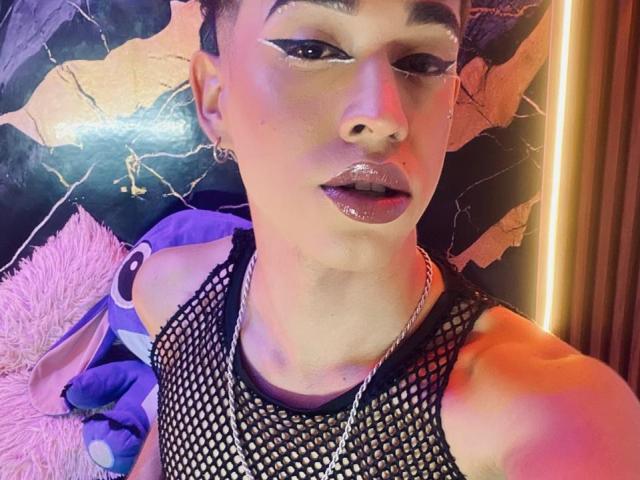 BriggitteLove with shiny lips, fishnet top, chain necklace, short hair, and bold eyeliner, looking at the camera. Pink fluffy pillow and purple blanket in the background.