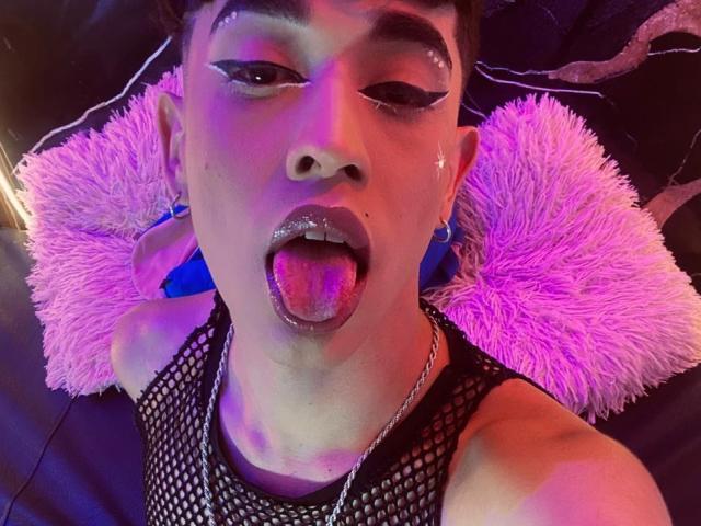 Close-up photo of BriggitteLove. They're lying on a dark surface with pink fluffy pillows. BriggitteLove has short, dark hair, thick black eyeliner, and a small star earring. Their tongue is out, coated in pink glitter. They're wearing a black fishnet top and a silver chain necklace. Purple and pink lighting creates a colorful effect. BriggitteLove has light skin and is making a playful, provocative expression. The photo is highly detailed and vibrant.