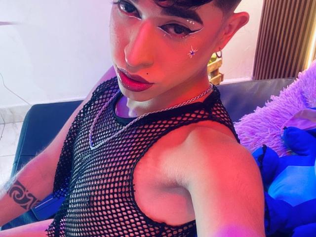 BriggitteLove with short curly brown hair, wearing a black fishnet top, red lipstick, and star-shaped eye makeup, showing a tattoo on their right arm. Pink and purple lighting.