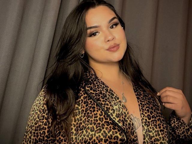 AnaSophia with long black hair, wearing a leopard print robe, showing a hint of her tits, smiling with brown eyes, wearing a silver necklace, and gray curtains in background.