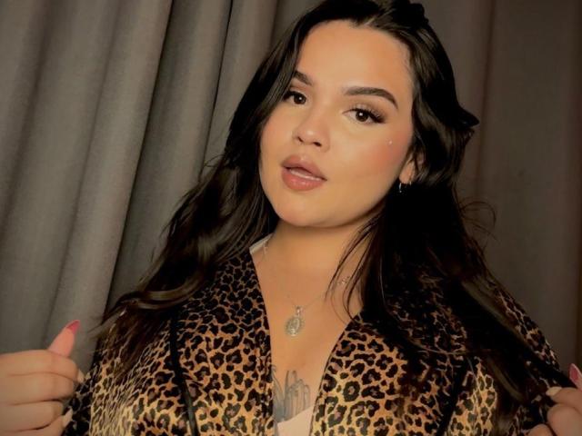 AnaSophia with long black hair, wearing a leopard print robe, has a cute face, pink lips, and a necklace. Her tits are covered by the robe. She's giving a thumbs-up.