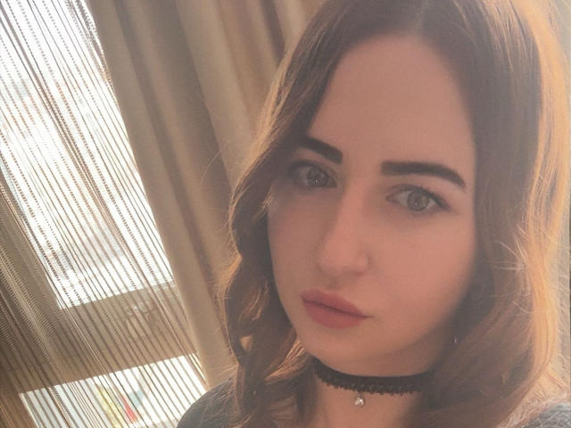 JulianaBella has light brown wavy hair, full eyebrows, pink lipstick, and a black choker with a small pendant. Her blue eyes look directly at the camera.