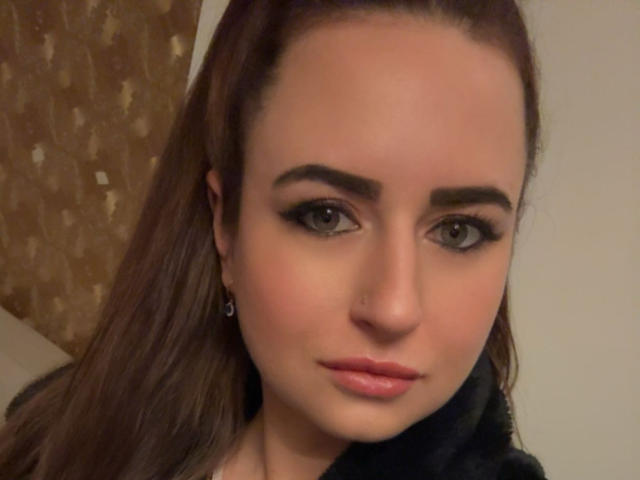 JulianaBella has brown eyes, thick black eyebrows, long brown hair, a small nose piercing, and glossy pink lips. Her makeup is dramatic with dark eyeliner.