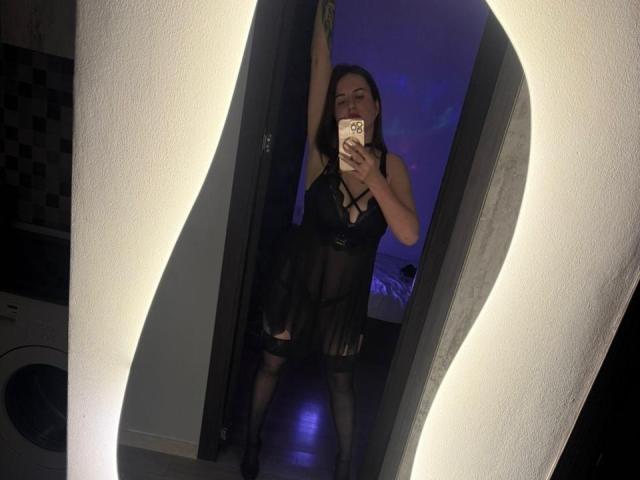 JulianaBella in black sheer lingerie, raising arm, showing her tits, standing in a wavy mirror, holding a phone, with a purple-lit background.