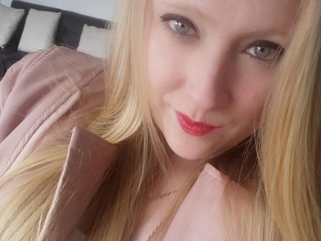 AliceCathyHot with blonde hair, light skin, and red lipstick, smiling at the camera with her blue eyes, wearing a pink jacket, and a thin gold necklace.