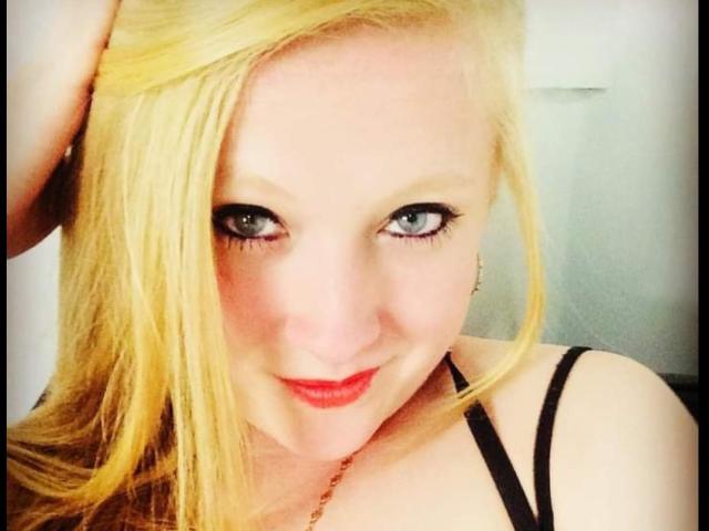 Close-up of AliceCathyHot with blonde hair, fair skin, and green eyes. She has black eyeliner, red lipstick, and is looking directly at the camera with a slight smile. Her right hand is in her hair. She's wearing a black strap top and a thin necklace. The image has a bright, soft focus with a slight blur in the background. Her makeup is bold, contrasting with her pale skin. The overall effect is cute and slightly seductive.