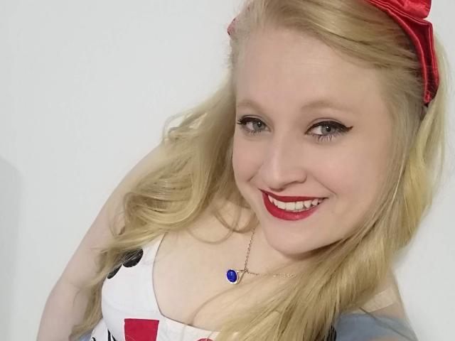 Close-up of AliceCathyHot, a blonde woman with wavy hair, a red headband, and bright red lipstick. She's wearing a white dress with black and red accents and a blue pendant necklace. She's smiling with slightly upturned lips, showing white teeth. Her eyes are light-colored, and she has subtle eyeliner. The image is well-lit, with a soft, even light. The composition focuses on her upper body and face.