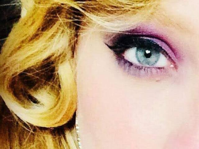 Close-up of AliceCathyHot's face showing a blue eye with heavy purple and pink eyeshadow, thick black eyeliner, and long lashes. Her wavy blonde hair is partially visible on the left. The eye is centered, with the eyeliner sharply contrasting against her light skin. The texture of the eyeshadow is smooth, and the eyeliner is precise. Bright light reflects in the eye.