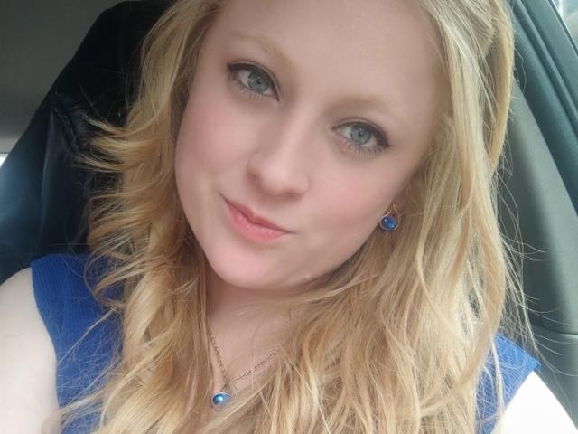 Close-up of AliceCathyHot with blonde wavy hair, light skin, and blue eyes. She wears a blue sleeveless top and small blue earrings with a gold chain. Her subtle makeup includes black eyeliner and pink lipstick. The image has a soft, natural light, highlighting her gentle smile. The composition centers on her face, with a slight upward angle. The texture of her hair is smooth, and her expression is calm and friendly.