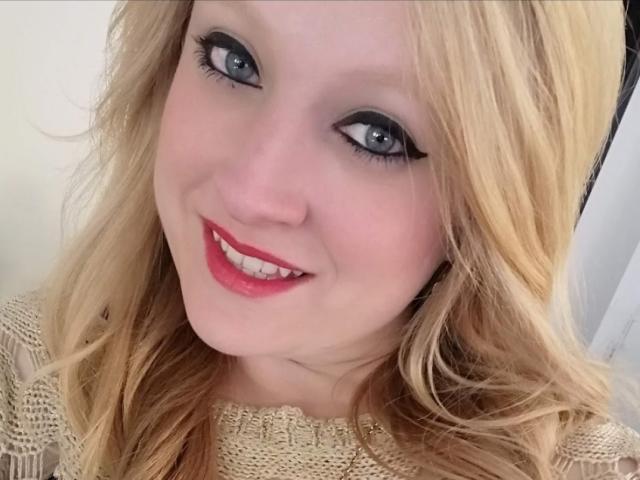 AliceCathyHot has blonde hair, blue eyes with black eyeliner, red lipstick, and a cute smile, wearing a beige knitted top, looking directly at the camera.