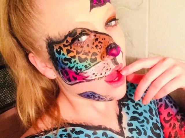 AliceCathyHot with leopard face paint, colorful spots, pink nose, and purple lips, touching her pink lip with her finger, wearing a leopard-print shirt. Blonde hair, blue eyes.
